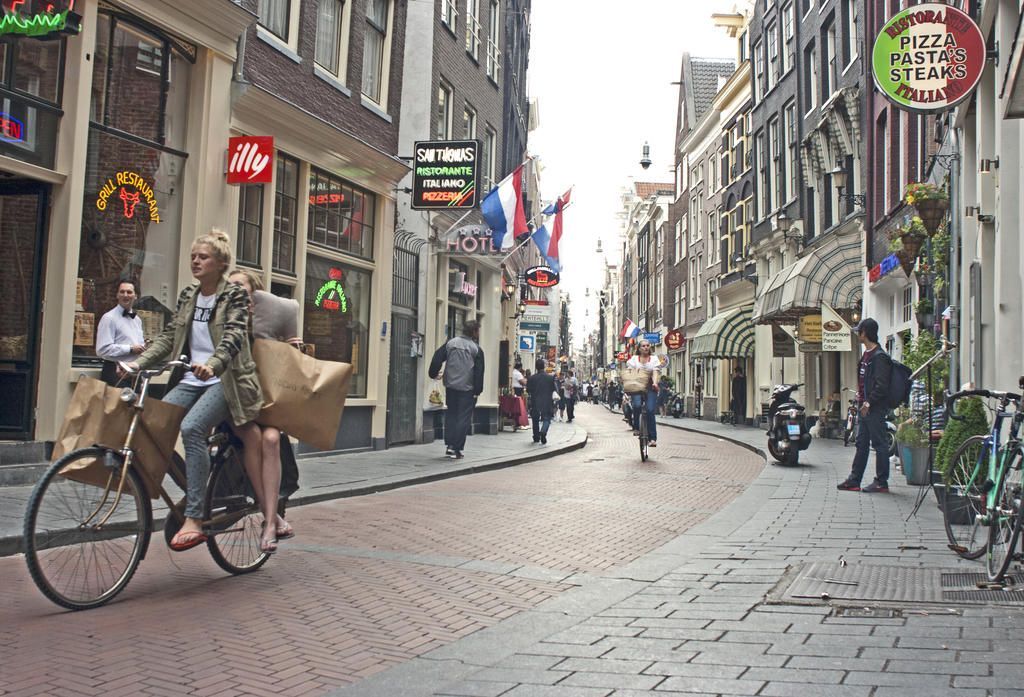 Cobblestone street in Amsterdam with cyclists, shops, and pedestrians. Buildings line the street, some with flags.