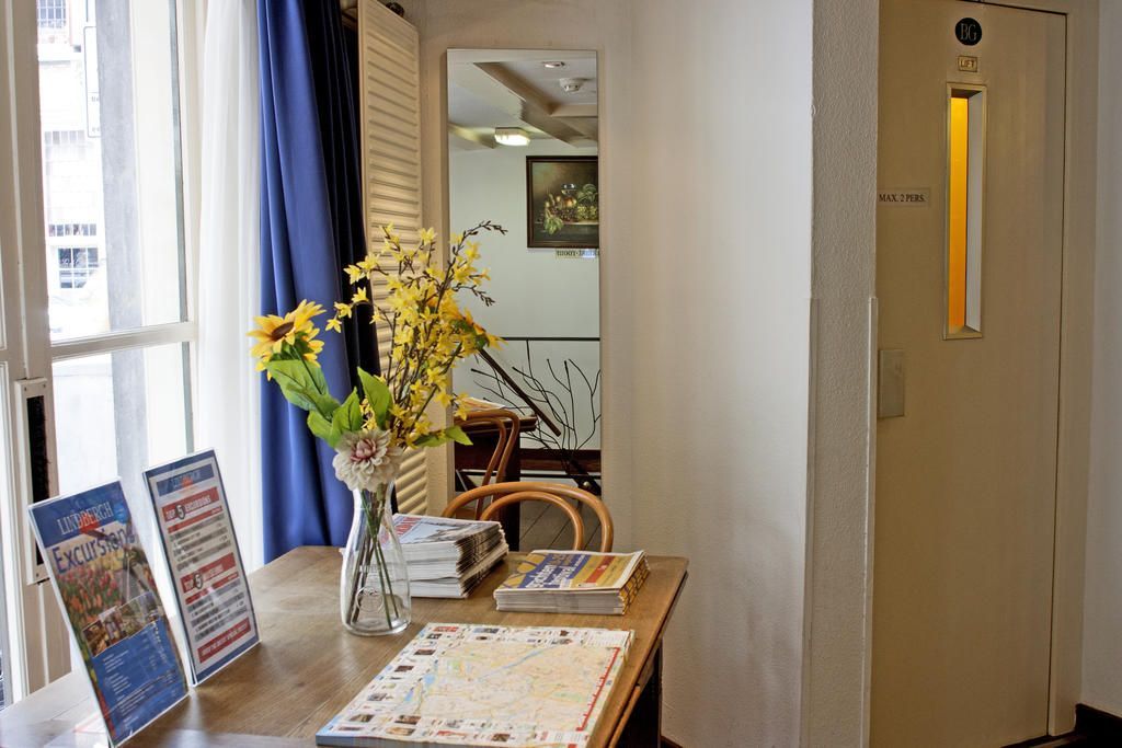 Interior with a wooden table, flowers, and brochures near a window and a white door. A mirror reflects stairs and a hallway.