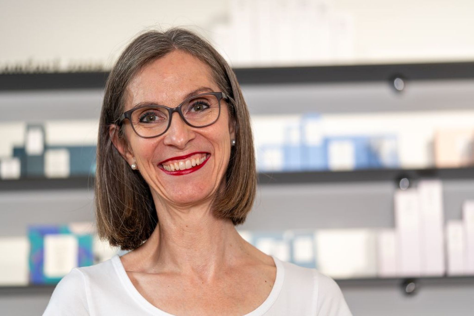 Eine Frau mit Brille und rotem Lippenstift lächelt vor einem Regal.