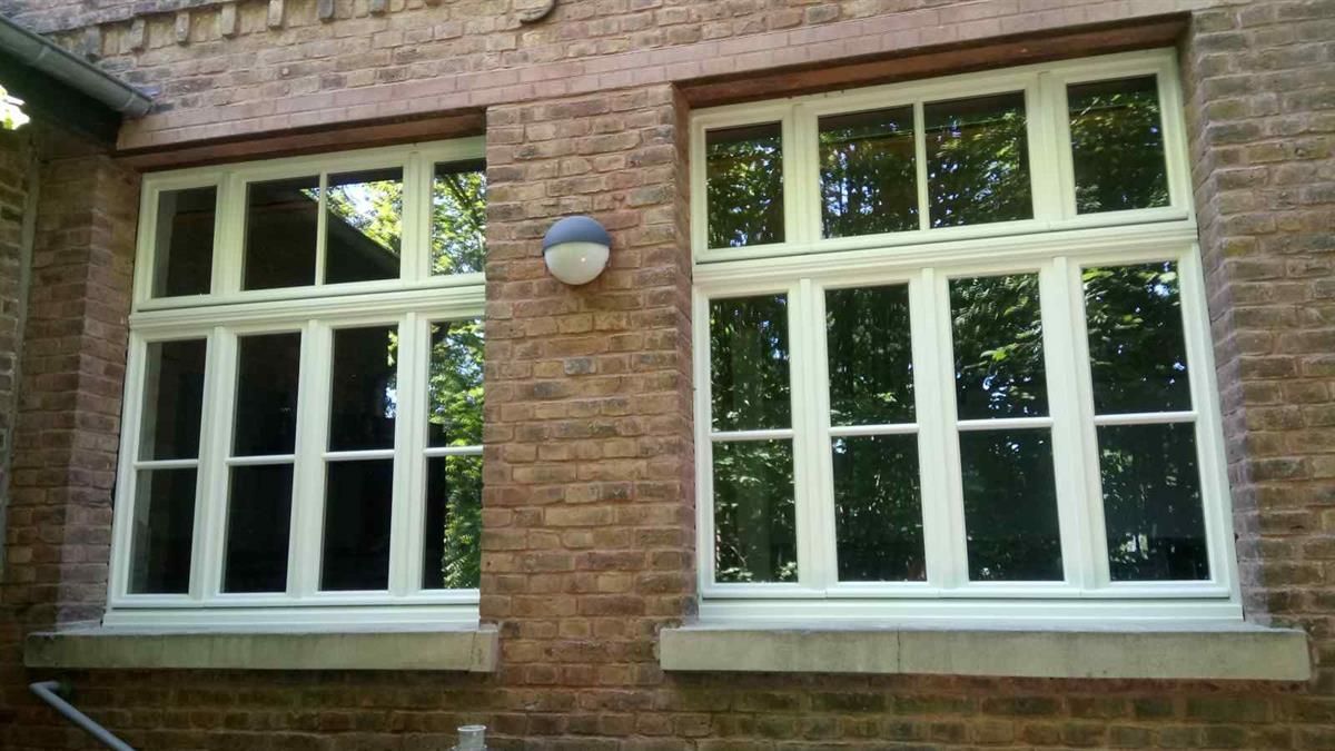 Zwei weiße Fenster an einem Backsteingebäude, in denen sich Bäume spiegeln