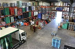 Interior de almacén con paletas, inventario apilado, carretillas elevadoras y un camión.