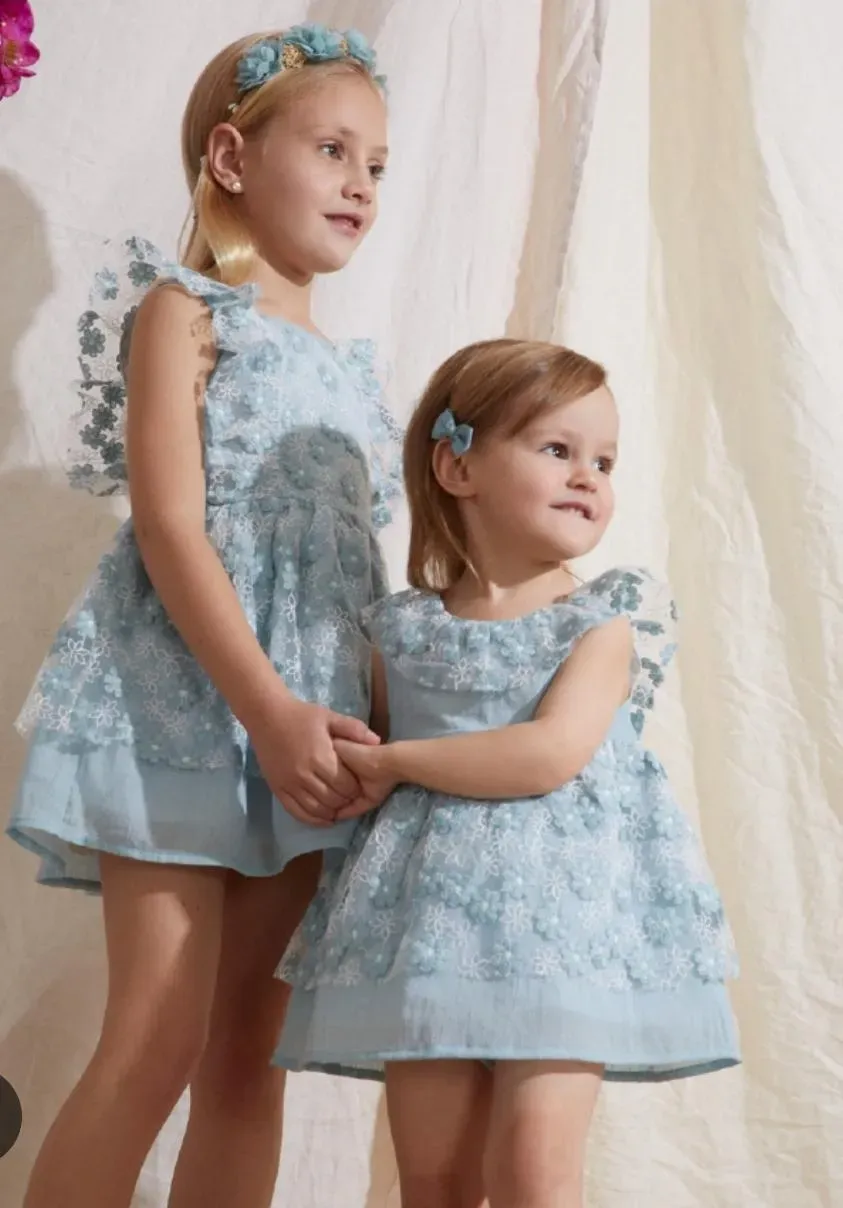 Dos niñas con vestidos azul claro a juego, de pie juntas en un entorno de estudio acogedor.