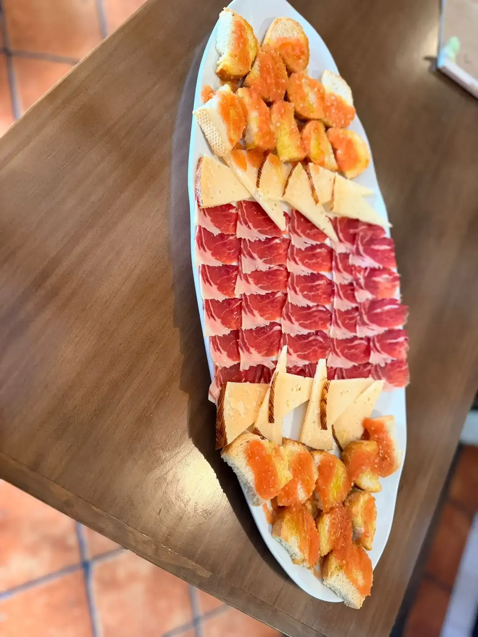 Variedad de frutas en rodajas y embutidos dispuestos en una larga bandeja blanca sobre una mesa de madera.