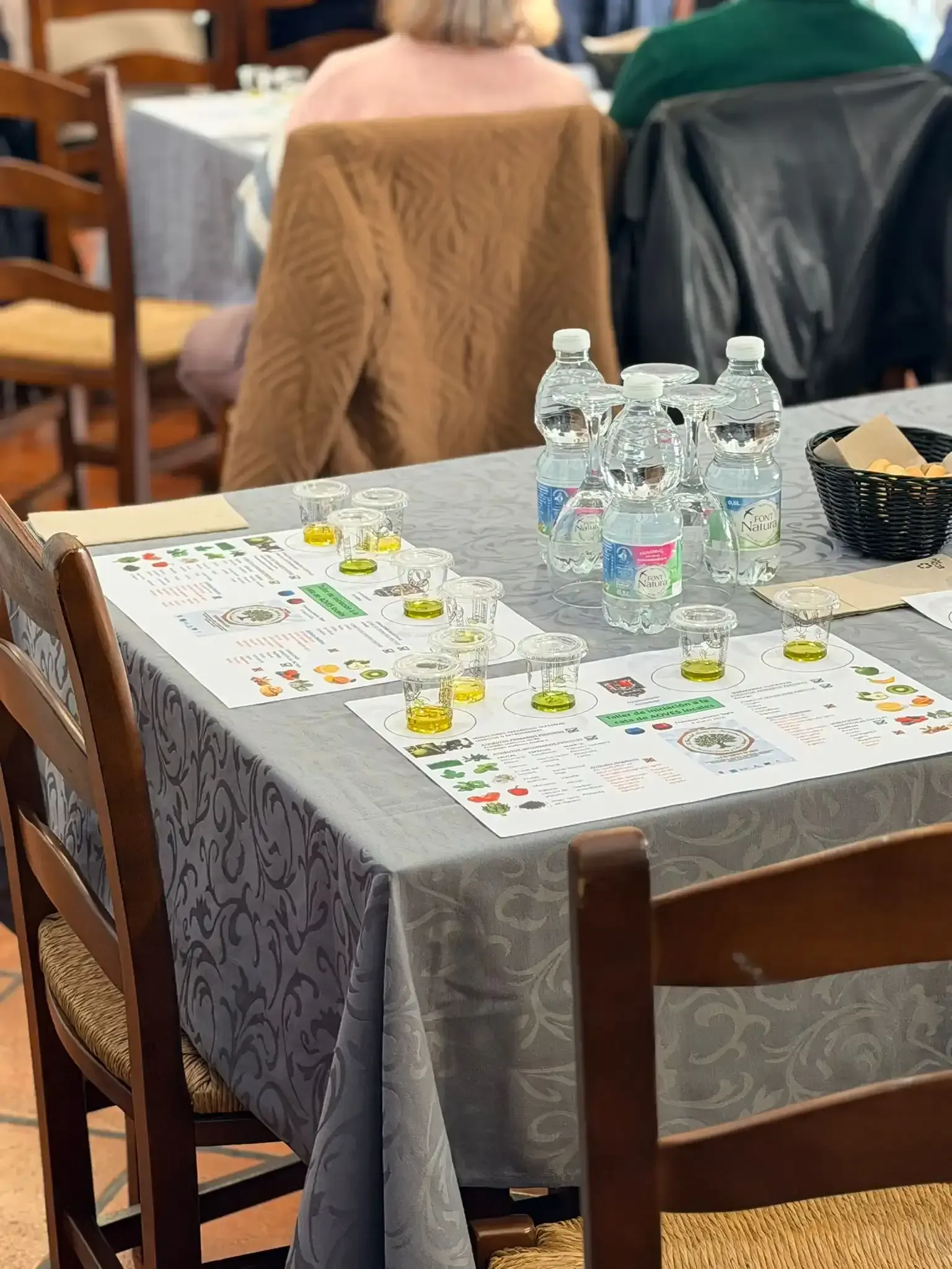 Mesa con mantel gris y agua embotellada en una cafetería, con sillas y gente sentada al fondo.