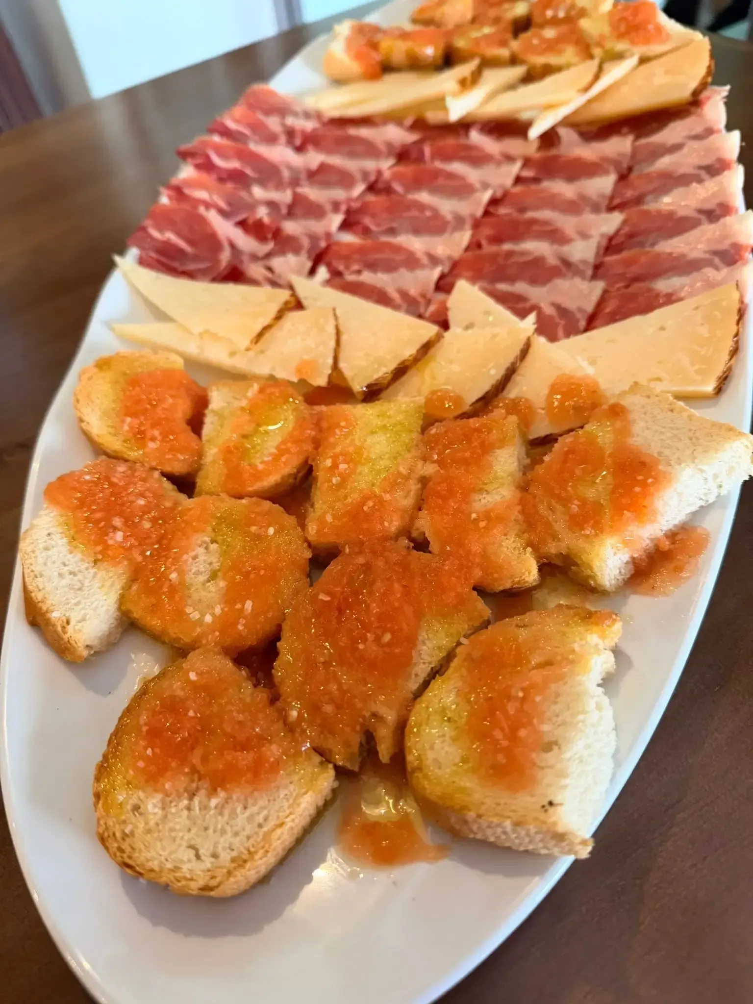 Variedad de embutidos y panecillos en una fuente blanca alargada, con aperitivos al fondo.