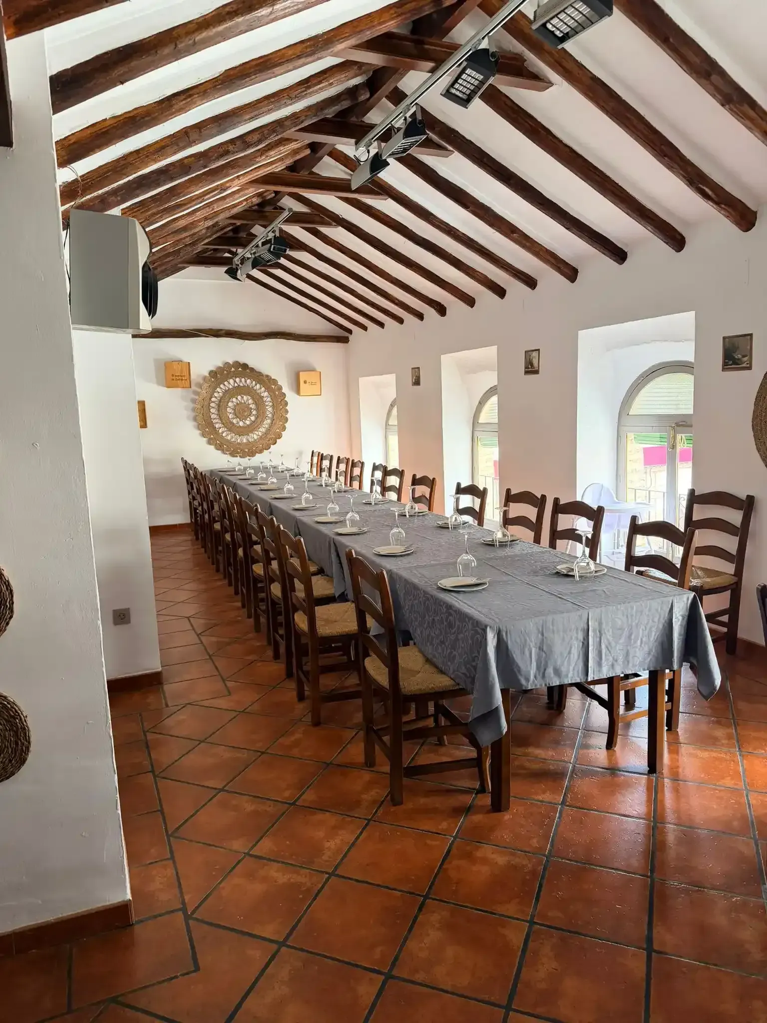 Amplio comedor con mesa puesta, sillas de madera, suelo de terracota y techo con vigas vistas.