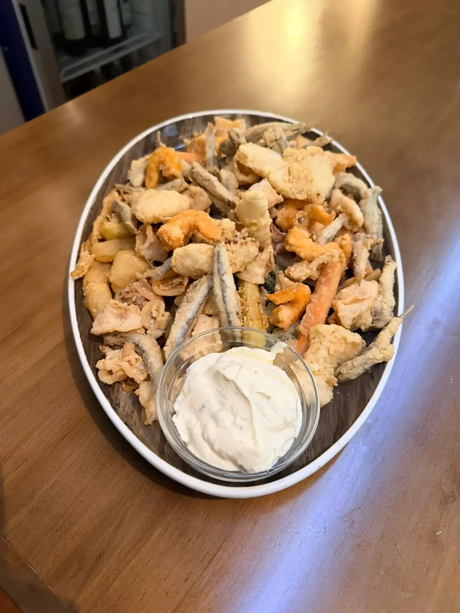 Bandeja de aperitivos fritos variados con una taza de salsa blanca para mojar sobre una mesa de madera.