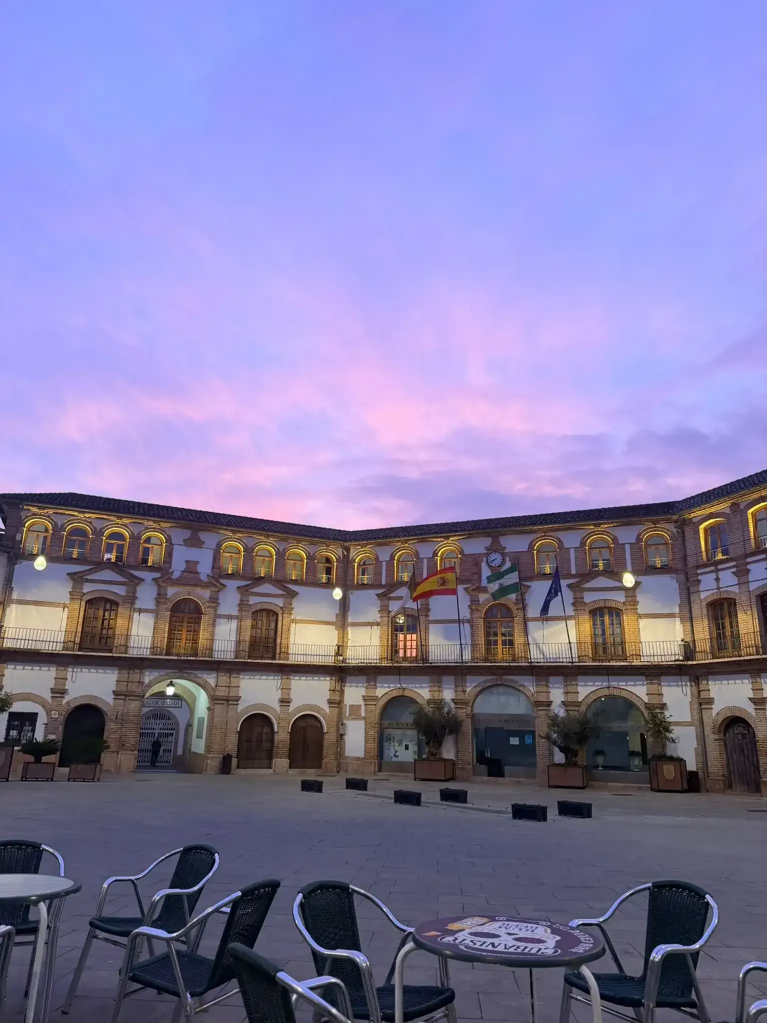 Patio de un edificio histórico con arcos al atardecer, con cielo rosa-púrpura y sillas de exterior en primer plano.