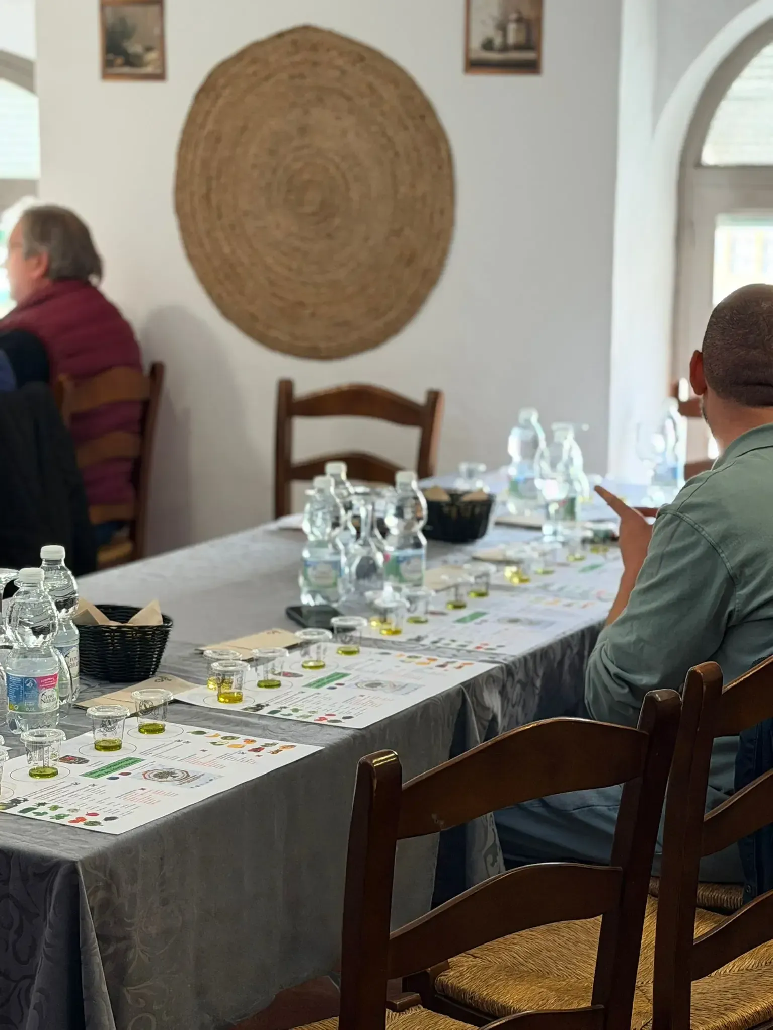 Dos personas están sentadas en una mesa larga con botellas de agua en una habitación luminosa con ventanas arqueadas.