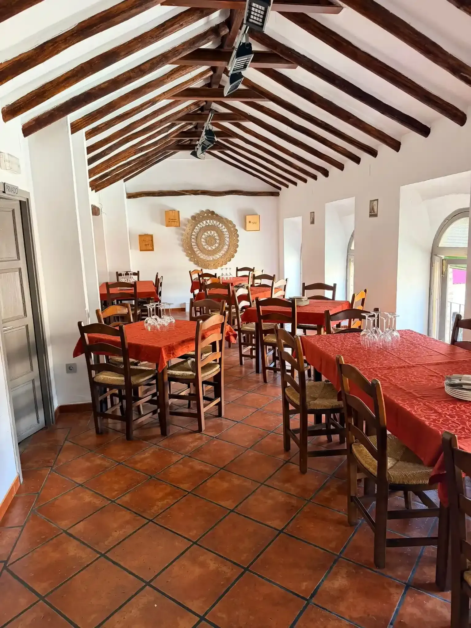 Comedor rústico vacío con mesas de madera, manteles rojos, suelo de terracota y techo con vigas vistas.