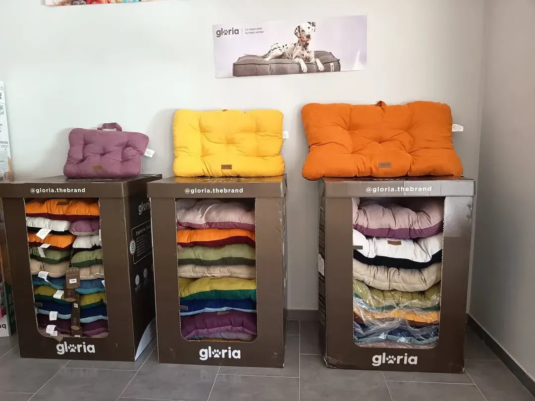 Exhibición de camas para perros en una tienda. Tres cajas exhiben camas plegables; encima, cojines de colores.