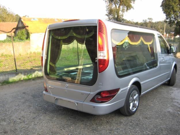 Carro funerário prateado estacionado em uma estrada de cascalho, com vidros fumê e cortinas decorativas.