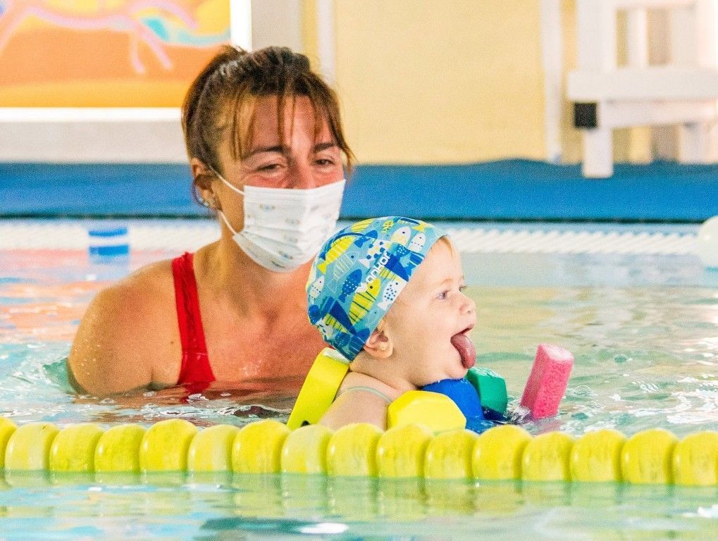 Instructor de natación enmascarado con un niño en la piscina usando un colorido chaleco salvavidas.