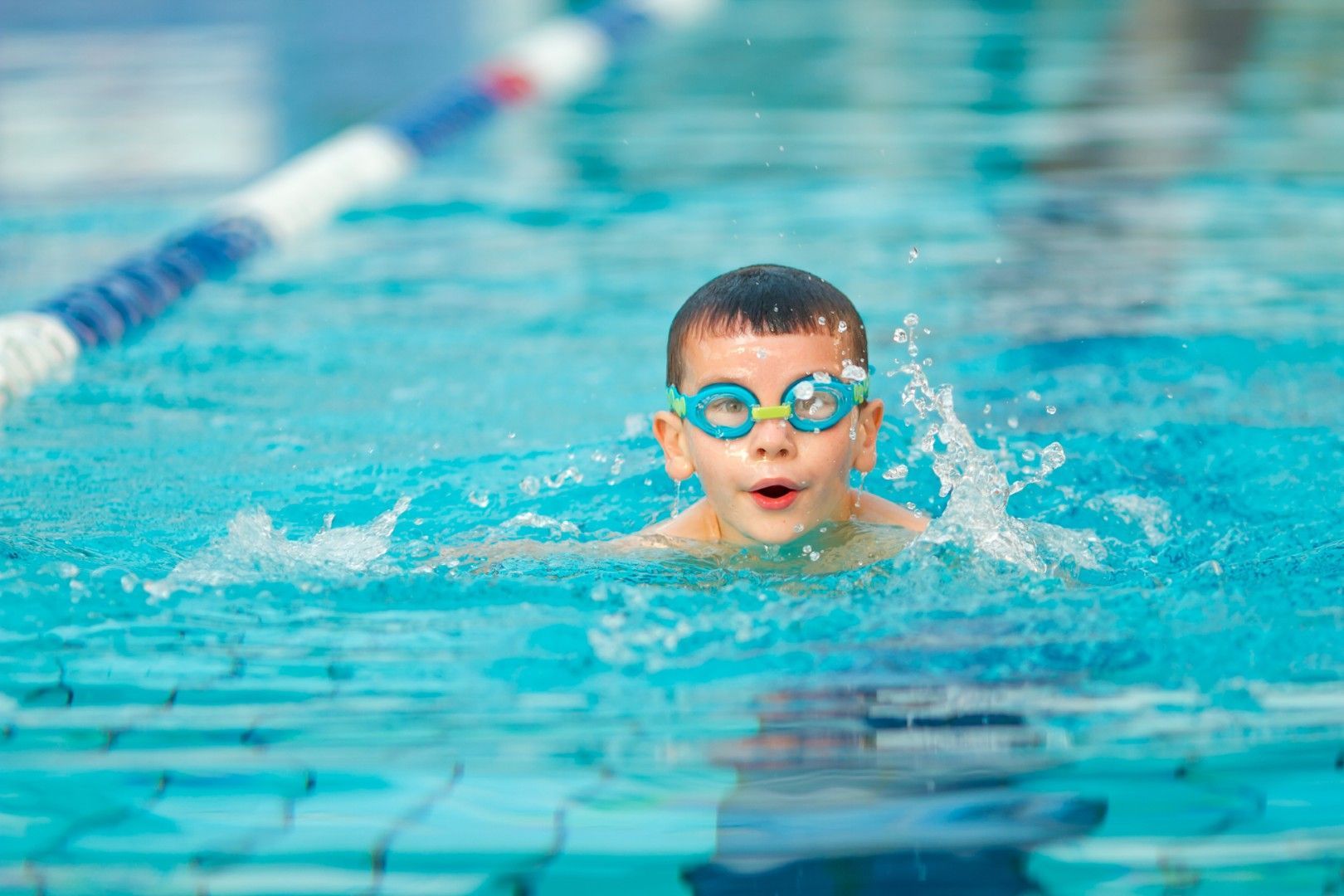 Un niño nada en una piscina de color turquesa, con gafas de natación azules, chapoteando hacia adelante.