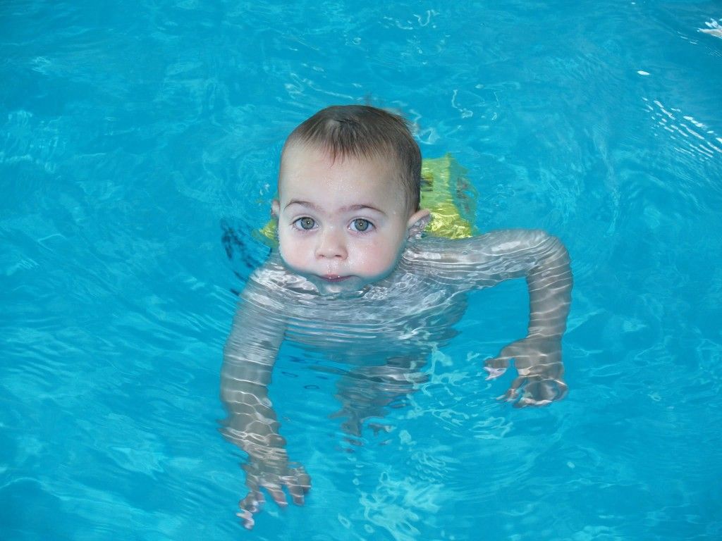 Un niño nada en una piscina de color azul brillante, mirando a la cámara con los brazos extendidos.