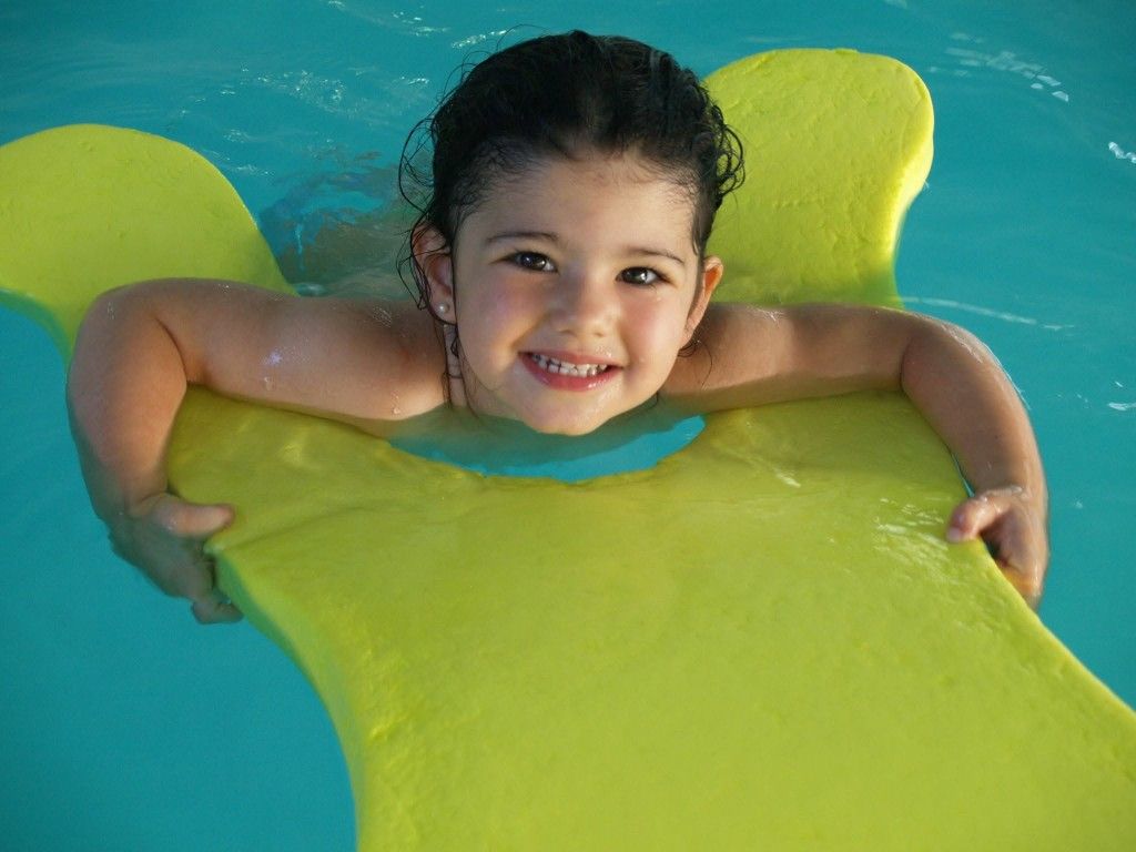 Un niño sonriente en una piscina, apoyado en un flotador de color verde brillante y mirando a la cámara.