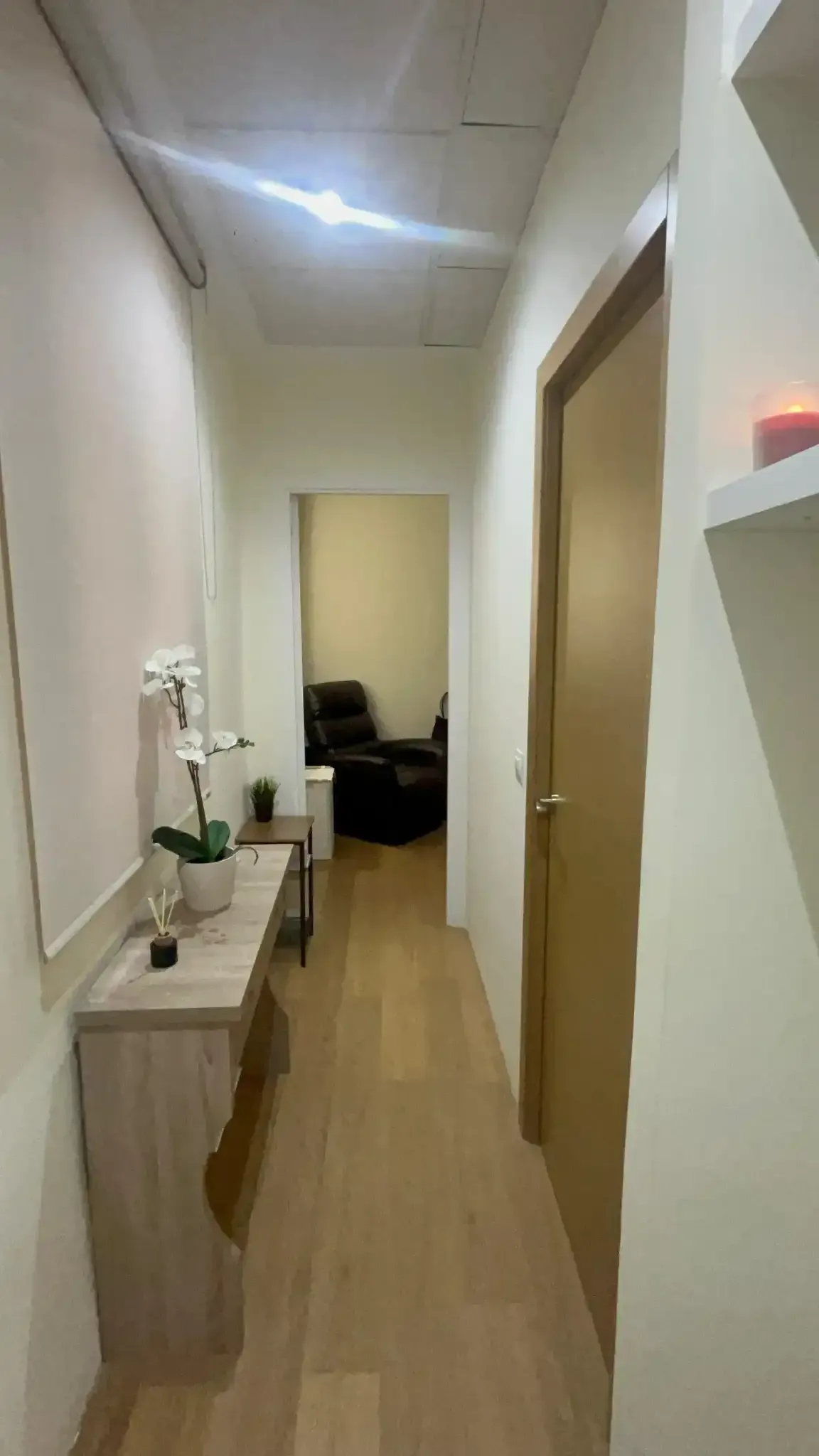 Pasillo estrecho con suelo de madera clara, mesa consola y puerta que conduce a una habitación con silla.