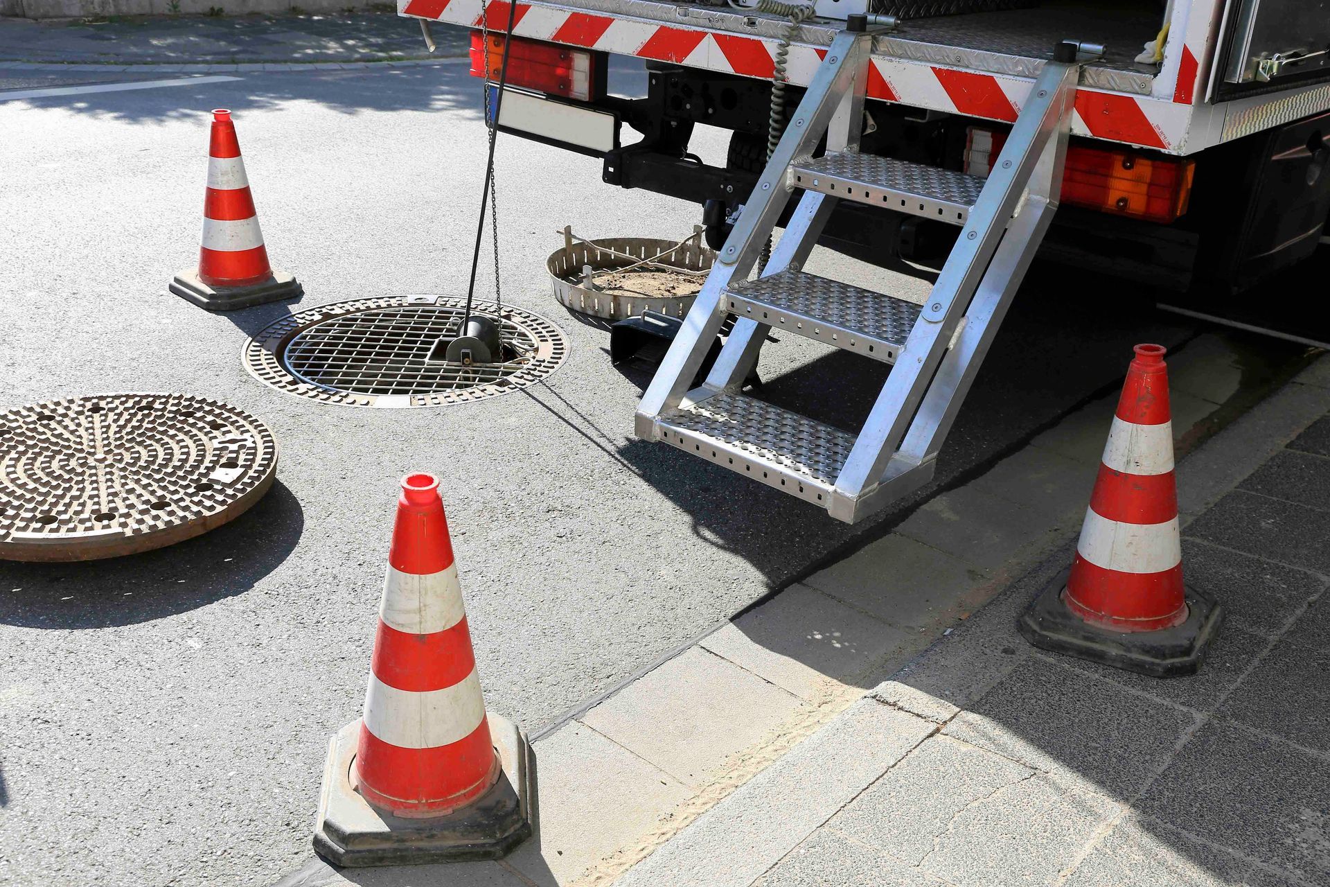 Straßenarbeiten: Schacht mit Leitkegeln und LKW geöffnet. Metallleiter heruntergelassen.
