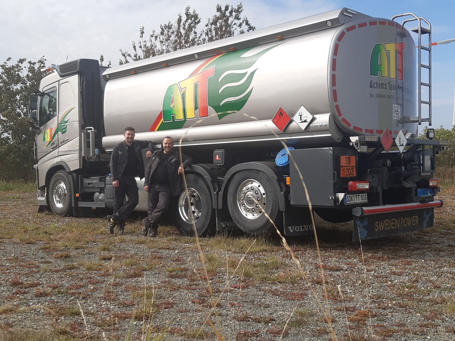 Zwei Männer stehen neben einem silbernen Tanklastwagen auf einer Rasenfläche; der Lastwagen hat ein grün-gelb-rotes Logo.