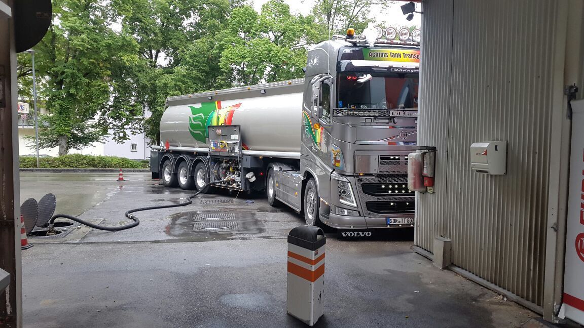 Silberner Tankwagen an einer Tankstelle, beim Befüllen. Nasser Asphalt, grüne und gelbe Akzente.