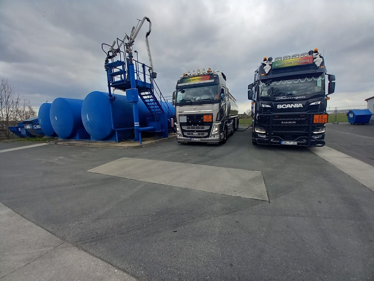 Zwei Tanklastwagen parken unter einem bewölkten Himmel neben blauen Lagertanks und einer Ladeplattform.