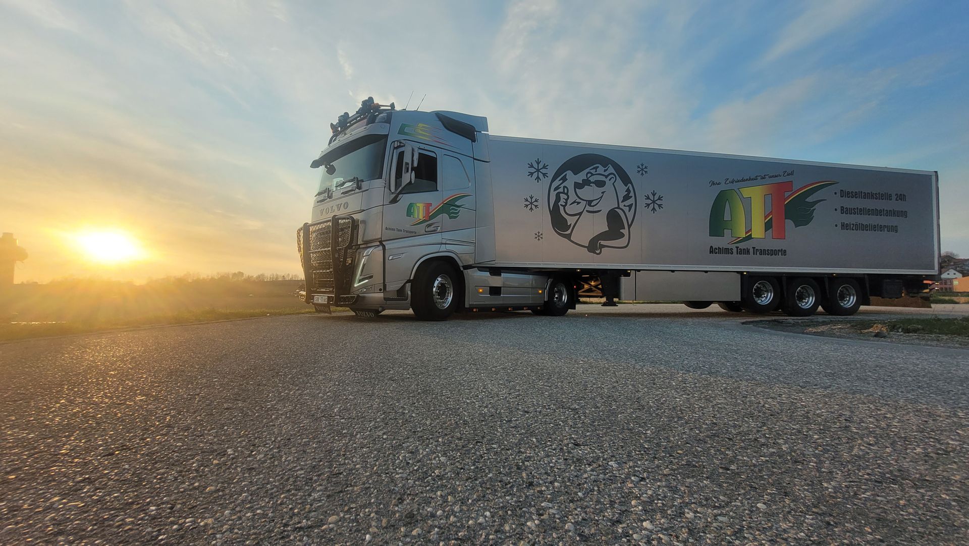 Silberner Sattelschlepper, der bei Sonnenuntergang auf Schotter geparkt ist. An der Seite befindet sich ein Logo mit der Grafik eines Gesichts und dem Wort „ATF“.