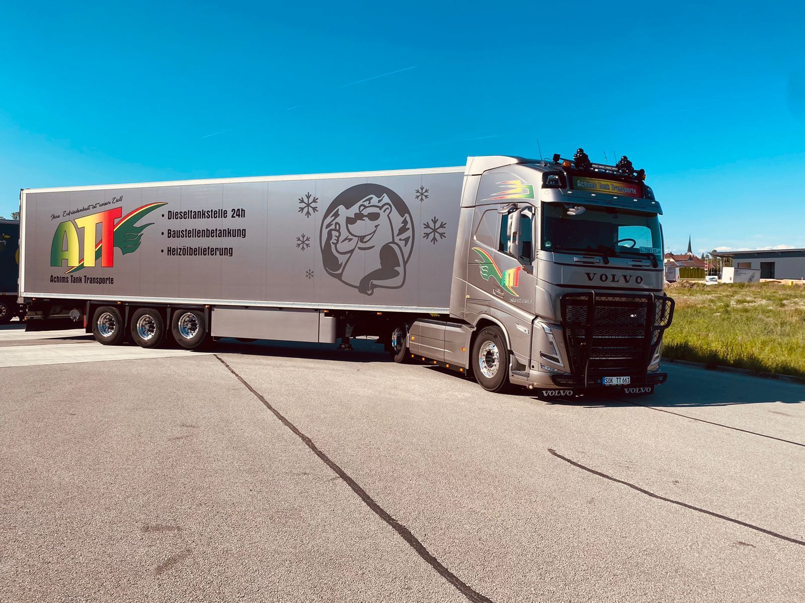 Silberner Sattelschlepper, geparkt auf einer grauen Fläche unter blauem Himmel. Der Anhänger hat ein Logo und eine Illustration.