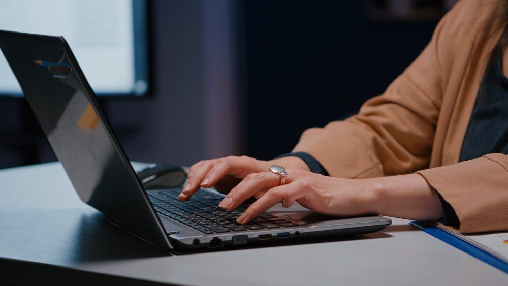 Una mujer está escribiendo en una computadora portátil en un escritorio.