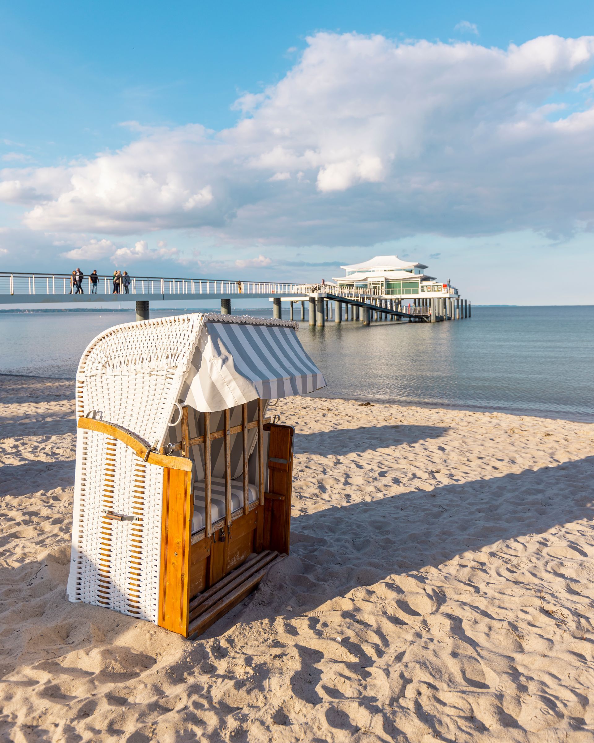 Strandkorb an einem Sandstrand mit einem Pier und bewölktem Himmel im Hintergrund.