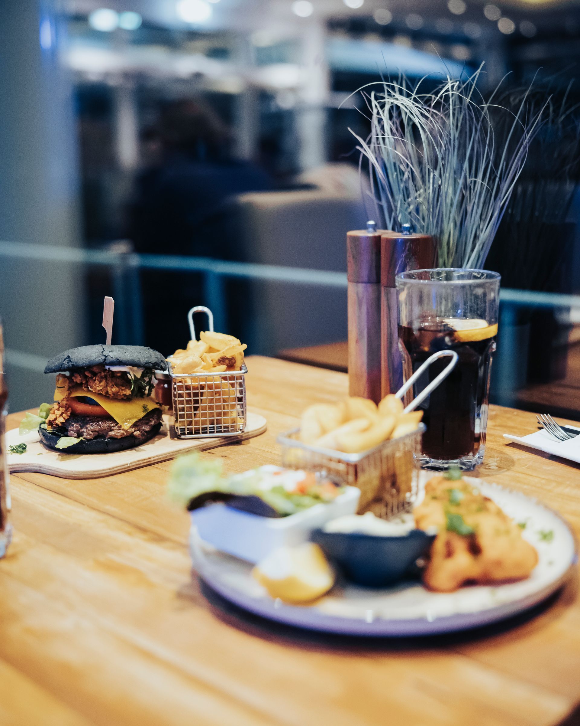 Ein Tisch mit einem schwarzen Burger, Pommes und einem Teller Essen, dazu ein Getränk. Die Kulisse ist ein Restaurant.