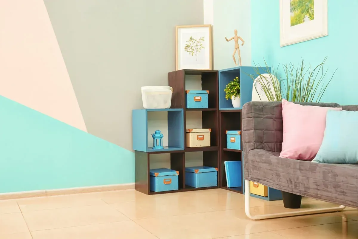 Pared geométrica en tonos pastel, una estantería de madera oscura con cajas y decoración azules, y un sofá gris con cojines.