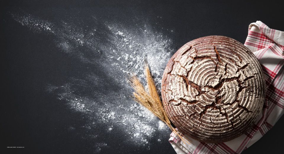 Ein runder Laib Brot mit rissiger Kruste liegt auf einer dunklen, mit Mehl bestäubten Oberfläche, daneben Weizenähren und ein kariertes Tuch.