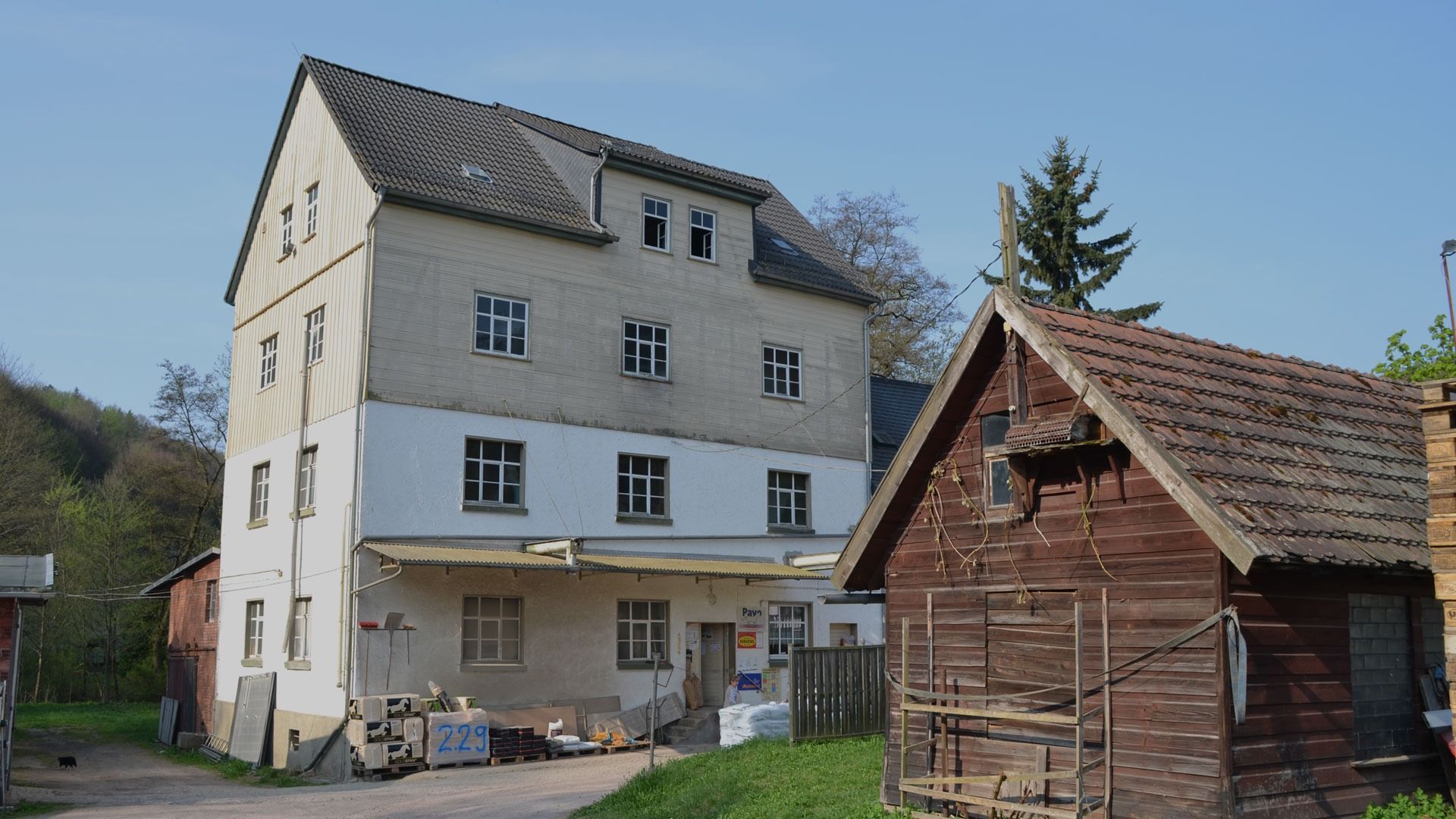 Ein mehrstöckiges Gebäude mit heller Fassade neben einem kleinen Holzschuppen, beide unter blauem Himmel.