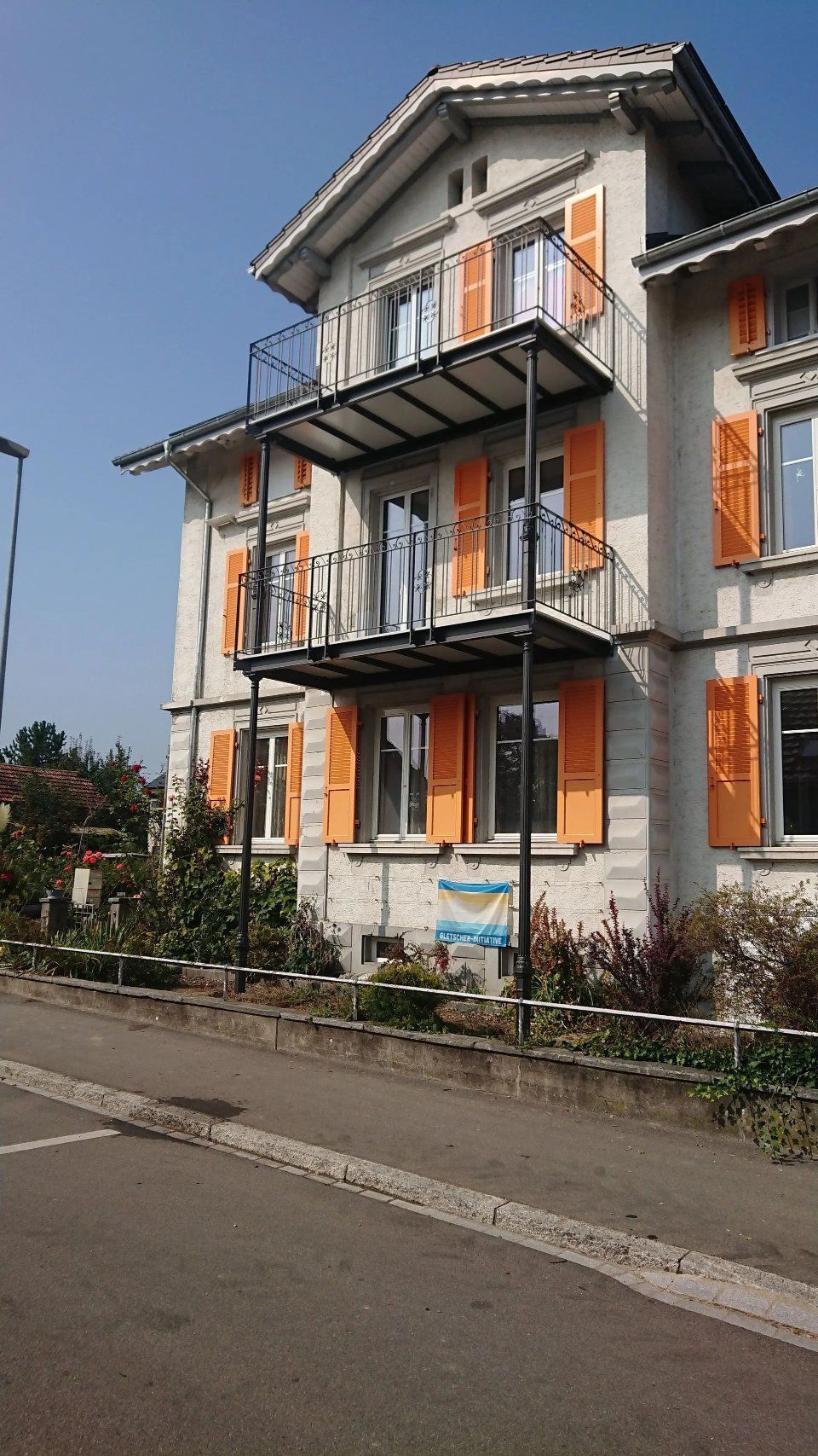 Dreistöckiges graues Haus mit orangefarbenen Fensterläden und Balkonen an einer Strasse unter klarem Himmel