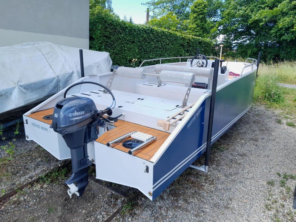 Kleines weiss-blaues Boot mit Aussenbordmotor, geparkt auf Kies neben einem überdachten Anhänger und Hecken.