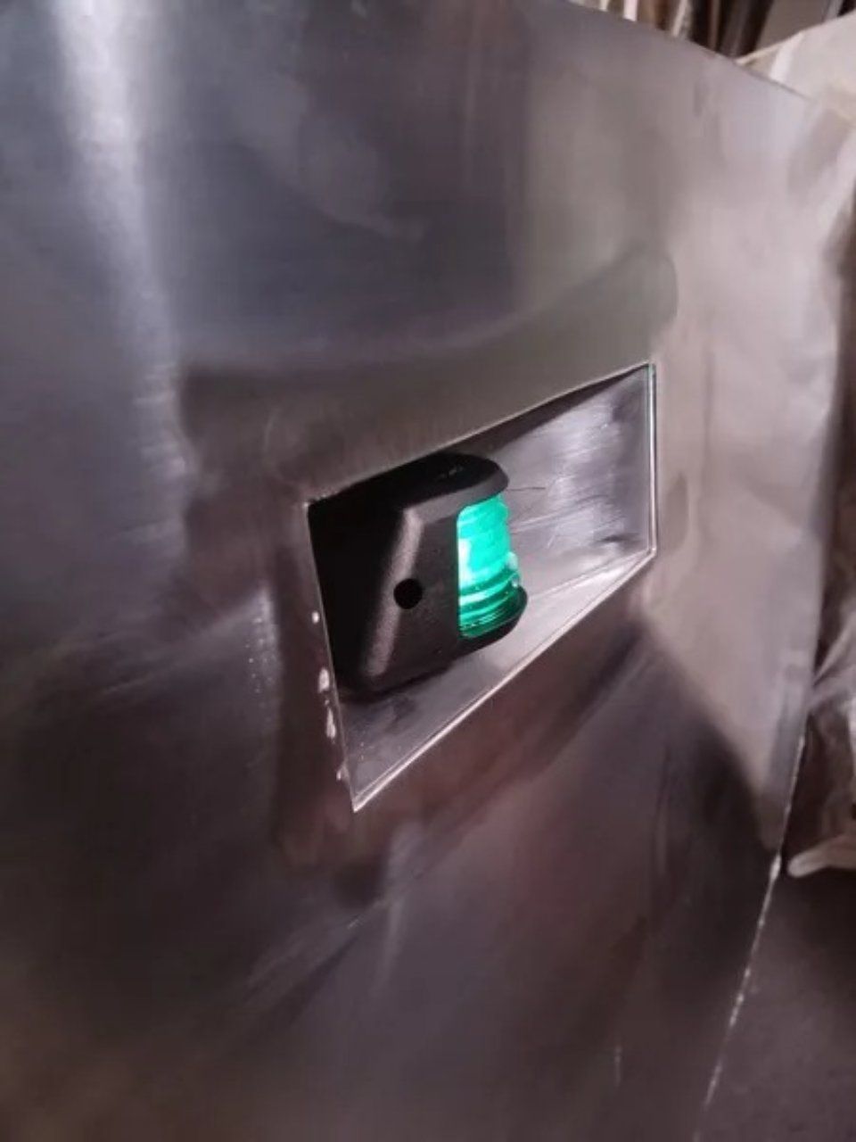 Nahaufnahme einer metallischen Wandplatte mit einem kleinen, grün beleuchteten Schalter oder einer Kontrollleuchte in einer rechteckigen Öffnung.