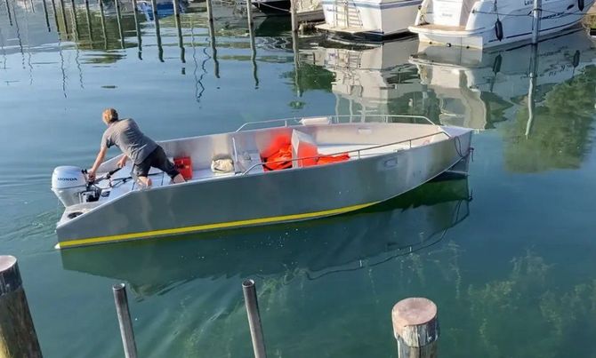 Kleines graues Motorboot mit zwei Personen, das auf ruhigem Hafenwasser in der Nähe von festgemachten Booten treibt.