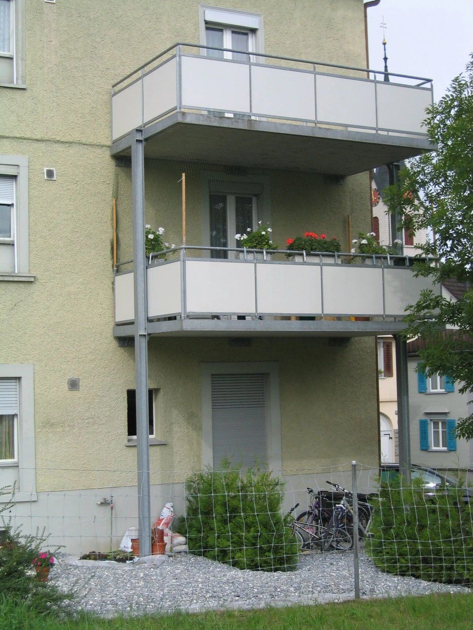 Balkon eines Mehrfamilienhauses mit Topfpflanzen und einem kleinen Garten darunter