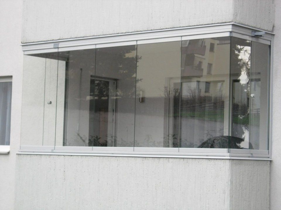 Ecke eines modernen Gebäudes mit einem grossen, verglasten Balkon und Spiegelungen in den Fenstern