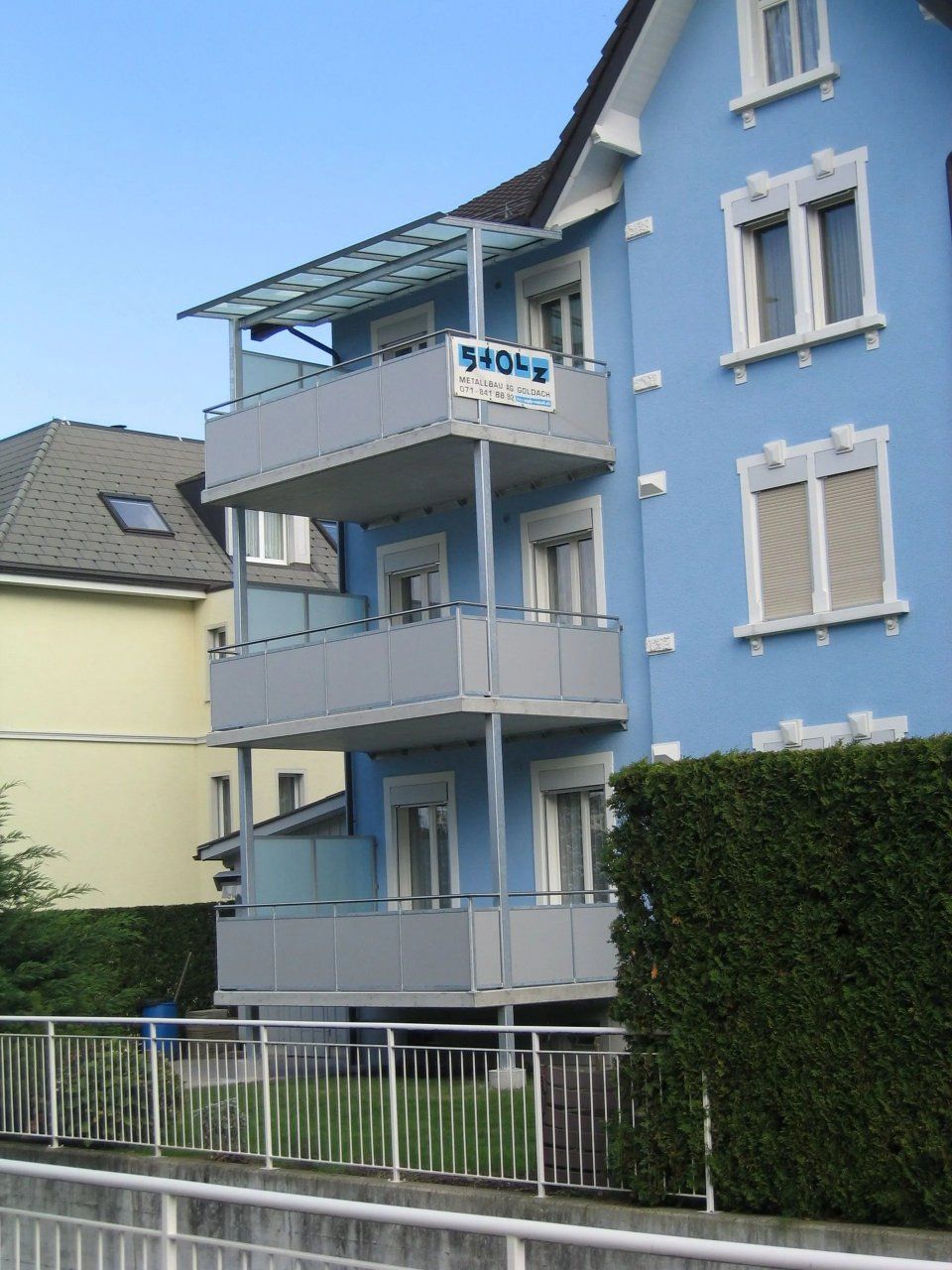 Blaues Wohnhaus mit Balkonen und einer Hecke an der Strasse