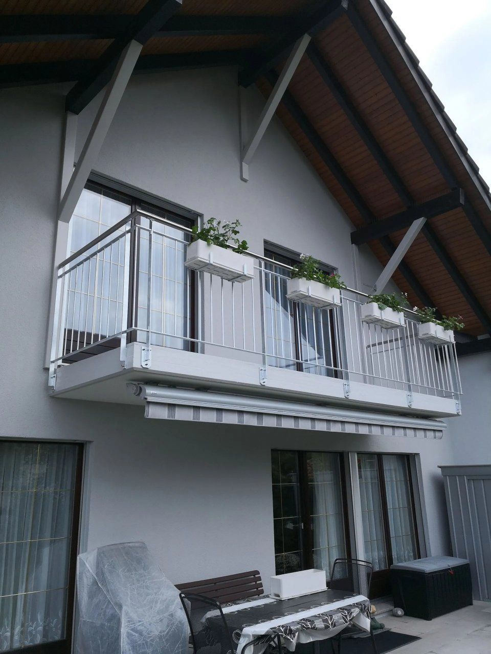 Balkon an einem modernen Haus mit weissem Geländer, Topfpflanzen und Glasschiebetüren unter einem schrägen Dach