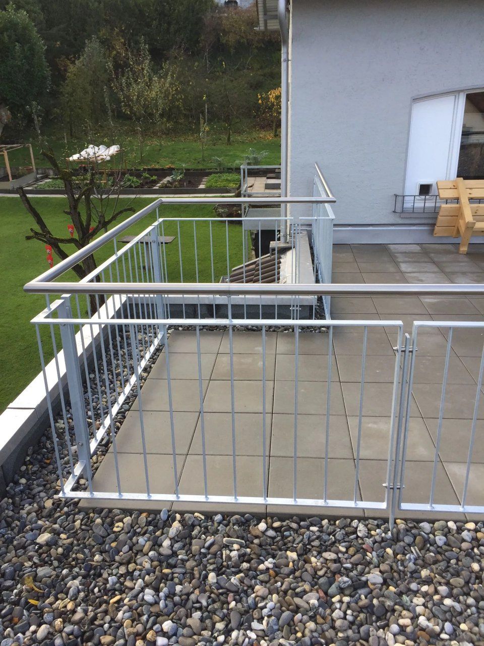 Moderne Terrasse mit weissem Geländer, grauen Pflastersteinen und Kies im Vordergrund, neben einem weissen Haus und Gartenblick