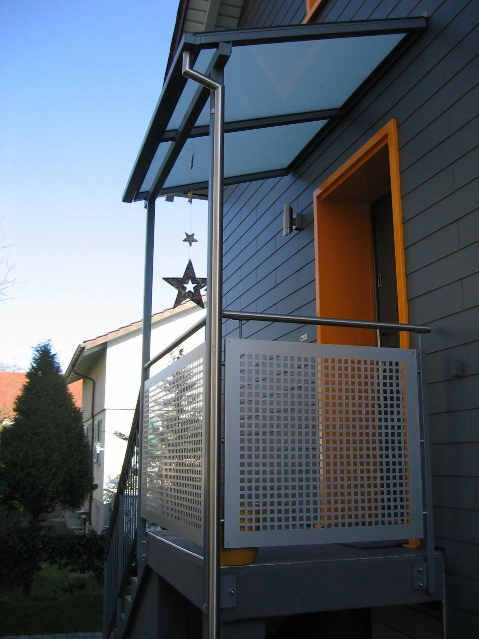 Kleine, moderne Veranda mit grauer Verkleidung, orangefarbener Tür und einem Stern als Dekoration unter einer Glasmarkise