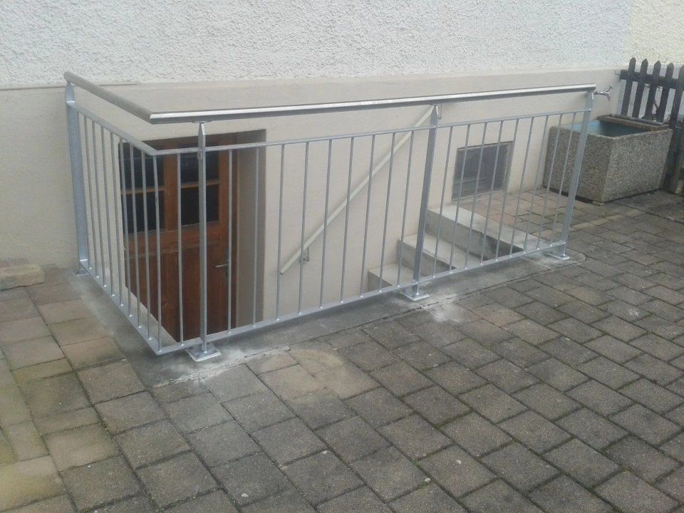 Metallgeländer umgibt ein Kellertreppenhaus neben einer gepflasterten Terrasse und einer weissen Wand.