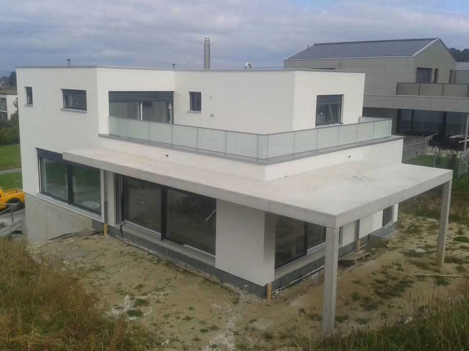 Modernes, weisses zweistöckiges Haus mit Glasbalkon und überdachter Terrasse auf einem sandigen Grundstück