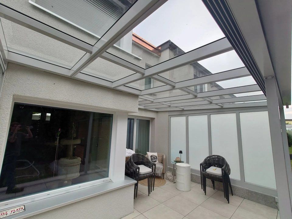 Überdachte Terrasse mit Glasdach, weissen Wänden und Terrassenstühlen neben Schiebetüren
