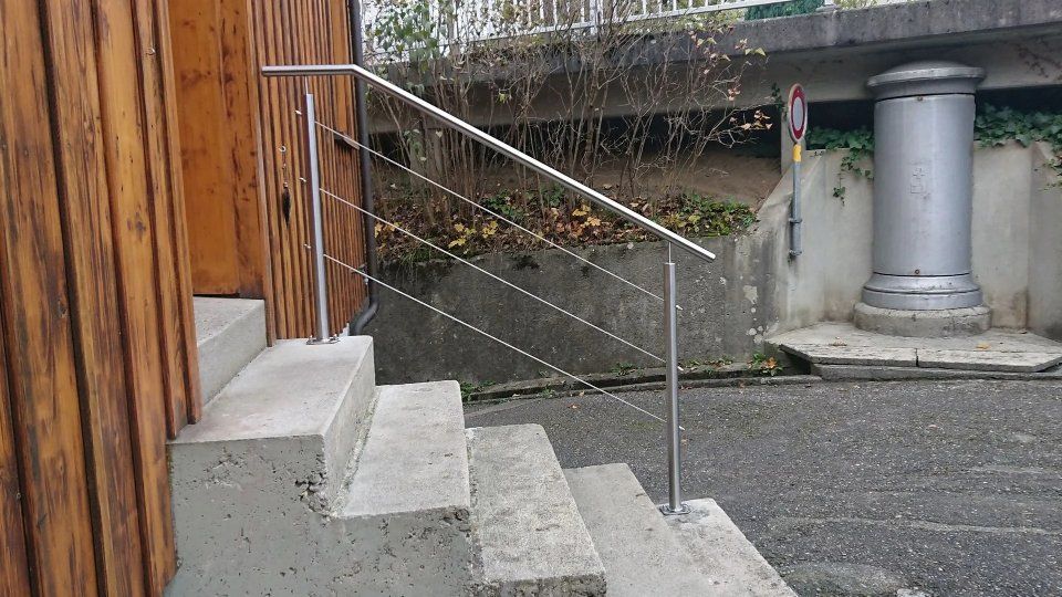 Eine Betontreppe mit Metallgeländer neben einem Holzgebäude führt zu einer Kiesfläche und einer grossen grauen Säule.