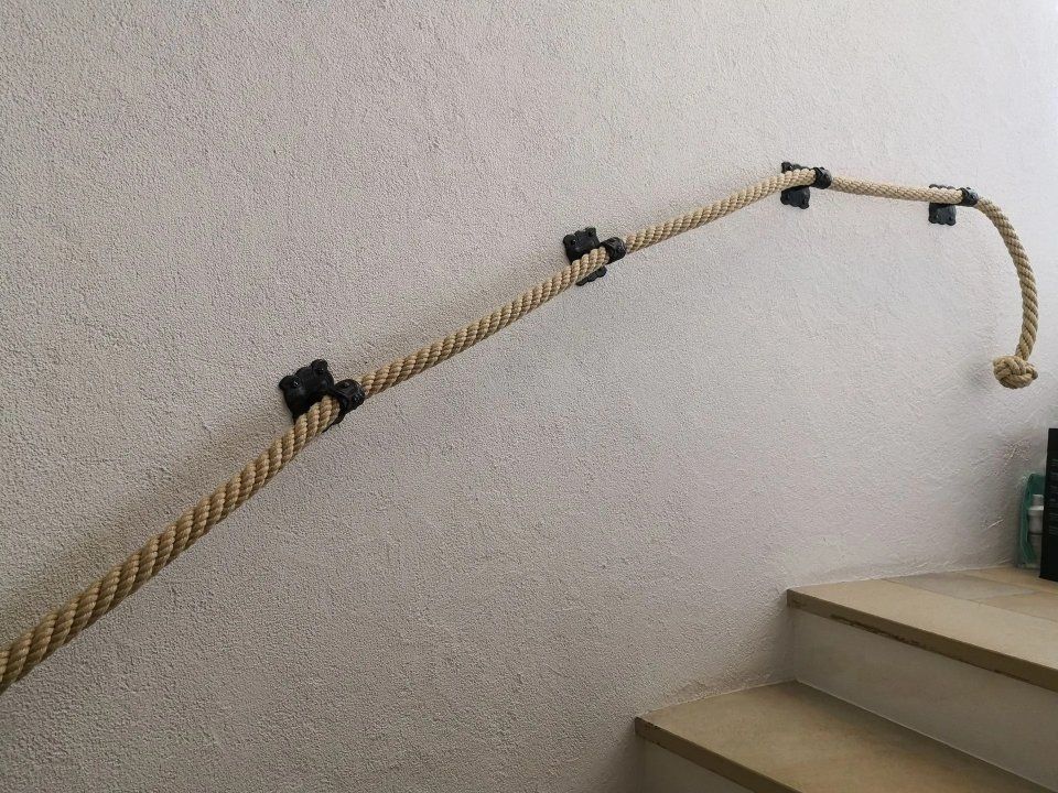 Ein geflochtenes Seilgeländer mit schwarzen Halterungen verläuft entlang einer Treppenhauswand, neben den mit Teppich ausgelegten Stufen.