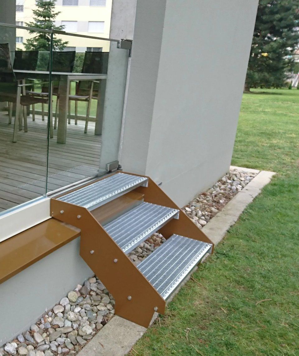 Metallstufen neben einem Gebäudeeingang, die von einer Rasenfläche und einem Kiesrand hinaufführen.