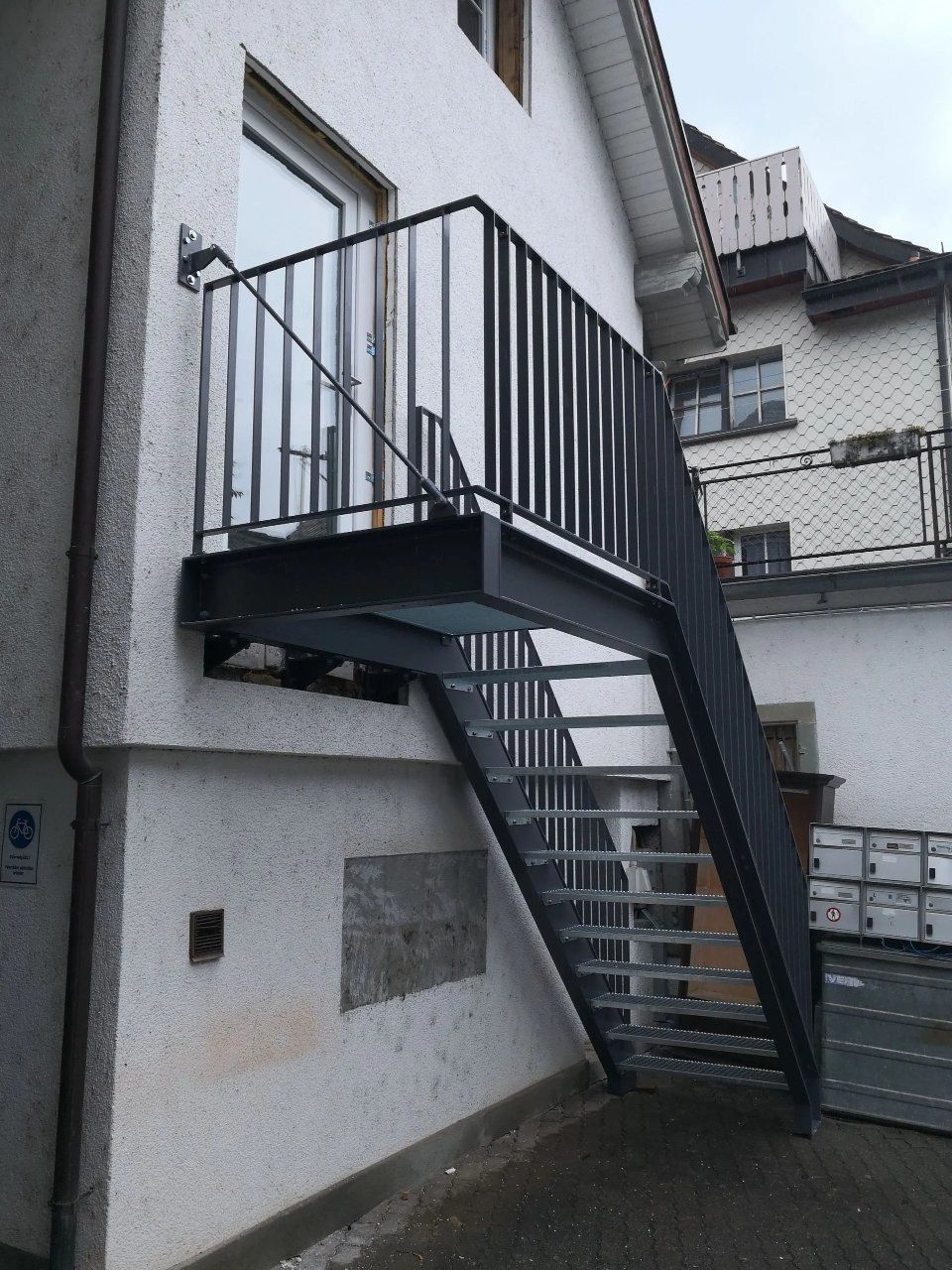 Eine Aussentreppe aus Metall und ein Podest sind an einer weissen Gebäudefassade angebracht.